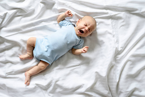 top view of baby in blue bodysuit and white pants lies yawning on white bed - Photo, Image
