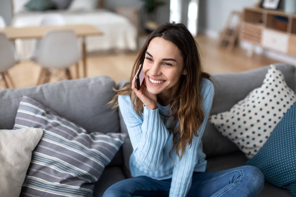 Attractive young woman talking on phone and smiling while sitting on a sofa at home. - Φωτογραφία, εικόνα