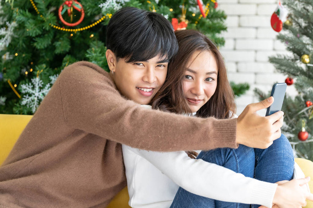 Young Asian adult teenager couple make their selfie photographing for celebrating Christmas holiday together in living room with Christmas tree decoration. - Zdjęcie, obraz