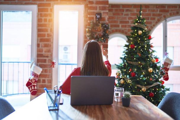 Hermosa mujer sentada en la mesa trabajando con el portátil en casa alrededor del árbol de Navidad posando hacia atrás apuntando hacia adelante con la mano del dedo
 - Foto, Imagen