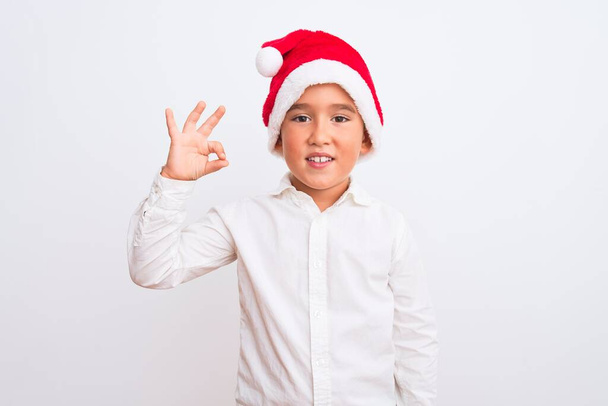 Hermoso niño con sombrero de Navidad Santa de pie sobre fondo blanco aislado sonriendo positivo haciendo signo de ok con la mano y los dedos. Expresión exitosa
. - Foto, imagen