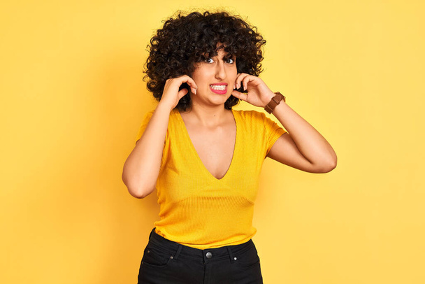 Młoda arabska kobieta z kręconymi włosami nosząca koszulkę stojącą nad odizolowanym żółtym tłem pokrywającą uszy palcem irytującym wyrazem na hałas głośnej muzyki. Koncepcja głuchoty. - Zdjęcie, obraz