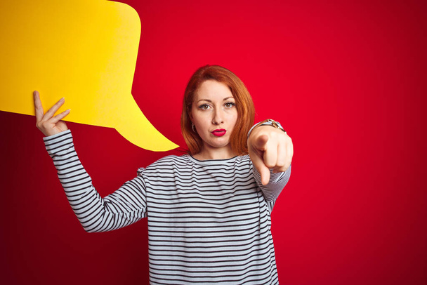 Young beautiful redhead woman holding yellow speech bubble over red isolated background pointing with finger to the camera and to you, hand sign, positive and confident gesture from the front - Photo, Image