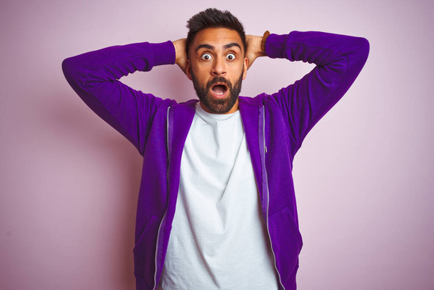 Young indian man wearing purple sweatshirt standing over isolated pink background Crazy and scared with hands on head, afraid and surprised of shock with open mouth - Photo, Image