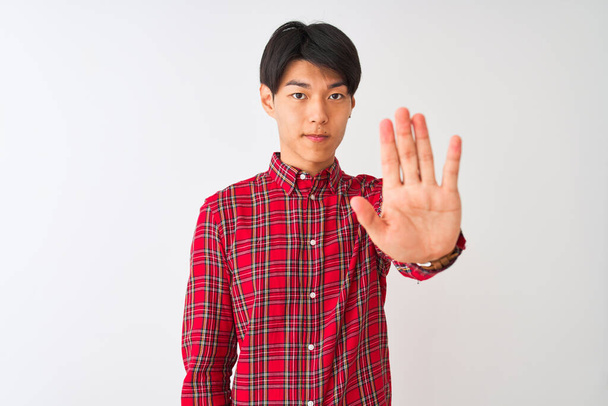 Joven hombre chino con camisa roja casual de pie sobre fondo blanco aislado haciendo dejar de cantar con la palma de la mano. Expresión de advertencia con gesto negativo y serio en la cara
. - Foto, imagen