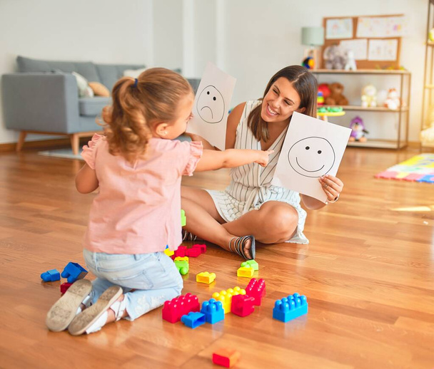 schöne Psychologin und blondes Kleinkind-Mädchen, das auf dem Boden sitzt und im Sprechzimmer eine Therapie mit Emoji-Emotionen macht - Foto, Bild