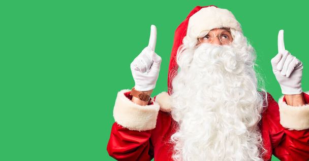Μεσήλικας όμορφος άνδρας φορώντας κοστούμι Άγιος Βασίλης και γενειάδα στέκεται χαμογελαστός κατάπληκτος και έκπληκτος και δείχνοντας επάνω με τα δάχτυλα και σήκωσε τα χέρια. - Φωτογραφία, εικόνα