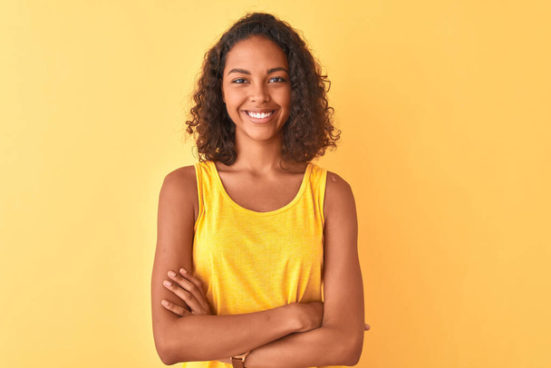 Jonge Braziliaanse vrouw die een t-shirt draagt en over een geïsoleerde gele achtergrond staat te glimlachen met gekruiste armen terwijl ze naar de camera kijkt. Positief persoon. - Foto, afbeelding