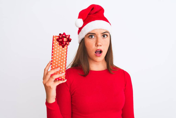 Νεαρό όμορφο κορίτσι φορώντας Χριστούγεννα Σάντα καπέλο κρατώντας δώρο πάνω από απομονωμένο λευκό φόντο φοβάται σε σοκ με ένα πρόσωπο έκπληξη, φοβισμένος και ενθουσιασμένος με την έκφραση του φόβου - Φωτογραφία, εικόνα