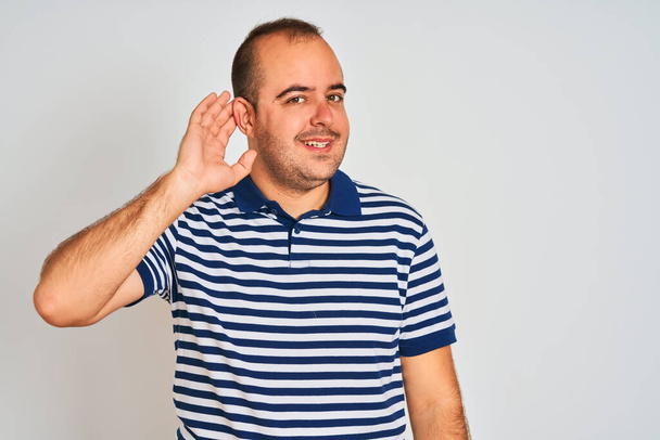 Молодий чоловік у повсякденному смугастому поло стоїть над ізольованим білим тлом, посміхаючись рукою на вухо слухаючи чутки або плітки. Концепція глухоти
. - Фото, зображення