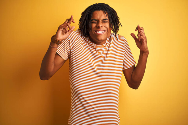 Afro man with dreadlocks wearing striped t-shirt standing over isolated yellow background gesturing finger crossed smiling with hope and eyes closed. Luck and superstitious concept. - Photo, Image