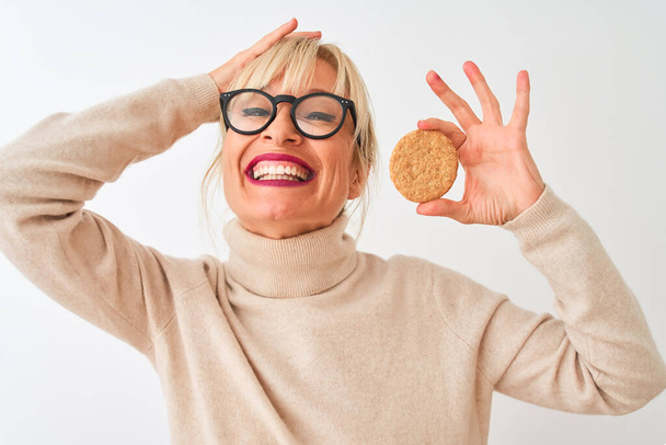Middle age woman wearing glasses holding cookie standing over isolated white background stressed with hand on head, shocked with shame and surprise face, angry and frustrated. Fear and upset for mistake. - Foto, Imagem