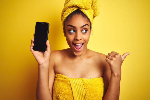 Afro mulher vestindo toalha após o chuveiro mostrando smatrphone sobre fundo amarelo isolado apontando e mostrando com o polegar até o lado com o rosto feliz sorrindo
 - Foto, Imagem