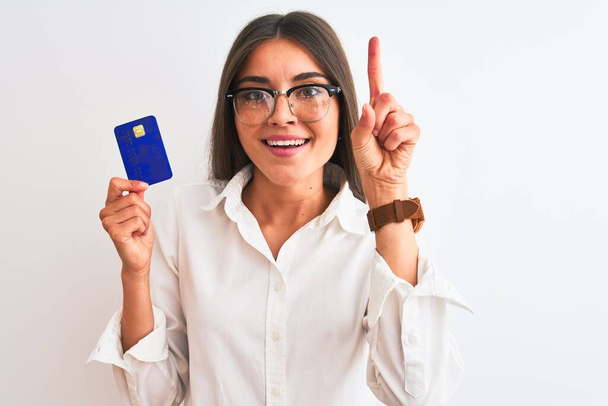 Gözlüklü genç bir iş kadını izole edilmiş beyaz arka planda kredi kartı tutuyor. Bir fikir ya da soru işareti ile şaşırıyor. - Fotoğraf, Görsel