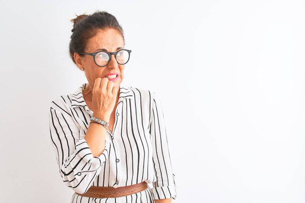 Podnikatelka středního věku nosí pruhované šaty a brýle na izolovaném bílém pozadí, vypadá vystresovaně a nervózně s rukama na ústech a kouše nehty. Úzkost problém. - Fotografie, Obrázek
