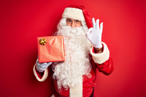 Noel Baba kostümü giyen orta yaşlı bir adam izole edilmiş kırmızı arka plan üzerinde hediyesini tutuyor. Tamam işareti yapıyor, mükemmel bir sembol. - Fotoğraf, Görsel