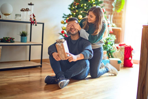 Mutlu ve kendinden emin gülümseyen genç bir çift. Erkeğin gözlerini kapayan bir kadın Noel ağacının etrafında hediyeyle dolaşır. - Fotoğraf, Görsel