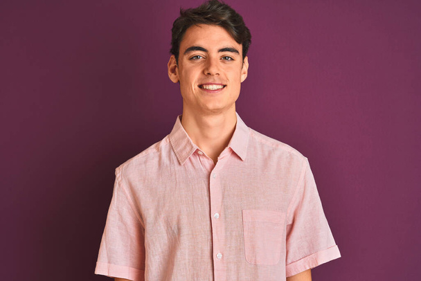 Мальчик-подросток в розовой рубашке стоит на фиолетовом изолированном фоне с счастливой и прохладной улыбкой на лице. Счастливчик
. - Фото, изображение
