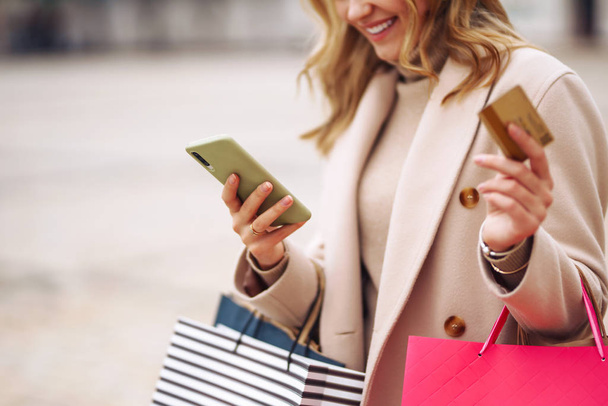Hands of beautiful woman with telephone and a credit card. Young woman holding shopping bags on the city street. Online shopping concept. Consumerism, purchases, shopping, lifestyle concept. - Foto, Imagen