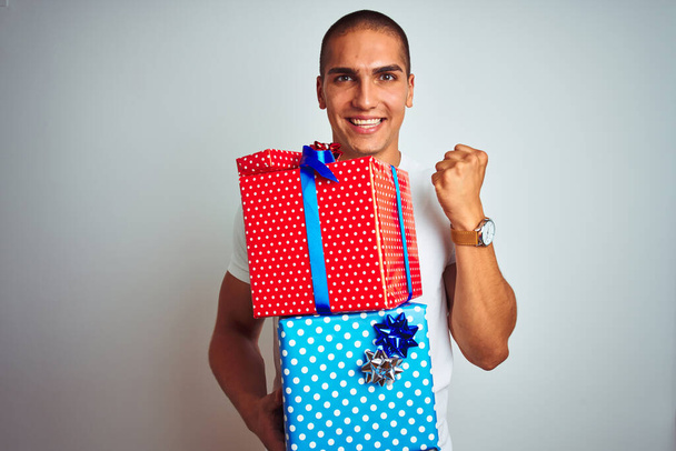 Fiatal jóképű férfi tartja születésnapi ajándékokat fehér elszigetelt háttér sikoltozó büszke és ünneplő győzelem és siker nagyon izgatott, éljenzés érzelem - Fotó, kép