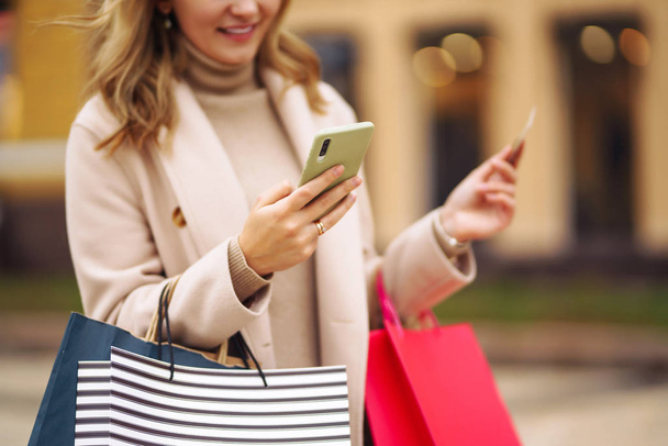 Hands of beautiful woman with telephone and a credit card. Young woman holding shopping bags on the city street. Online shopping concept. Consumerism, purchases, shopping, lifestyle concept. - Фото, изображение