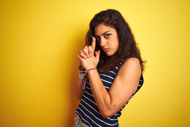 Mladá krásná žena v pruhovaném tričku, stojící nad izolovaným žlutým pozadím, držící symbolickou zbraň gestem ruky, hrající si na zabíjející střelné zbraně, rozzlobený obličej - Fotografie, Obrázek