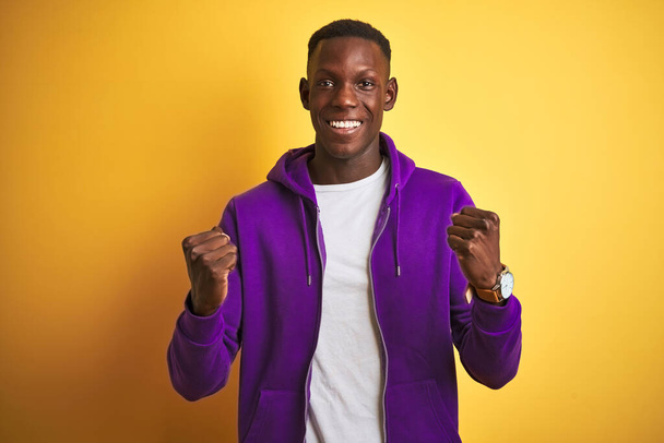 African american man wearing purple sweatshirt standing over isolated yellow background celebrating surprised and amazed for success with arms raised and open eyes. Winner concept. - Photo, Image