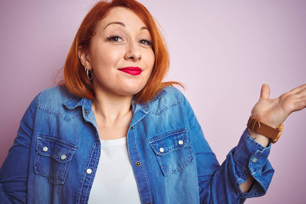Youg beautiful redhead woman wearing denim shirt standing over isolated pink background clueless and confused expression with arms and hands raised. Doubt concept. - Zdjęcie, obraz
