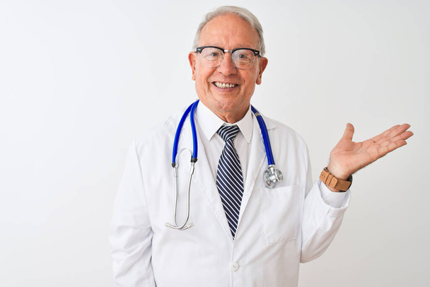 Senior grijs-harige arts draagt stethoscoop staan over geïsoleerde witte achtergrond lachende vrolijke presenteren en wijzen met palm van de hand kijken naar de camera. - Foto, afbeelding