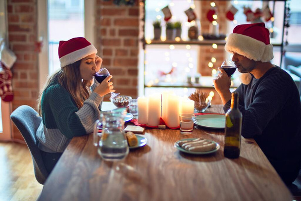 Jovem casal bonito sorrindo feliz e confiante. Comer comida e beber xícara de vinho usando chapéu de Papai Noel celebrando o Natal em casa
 - Foto, Imagem