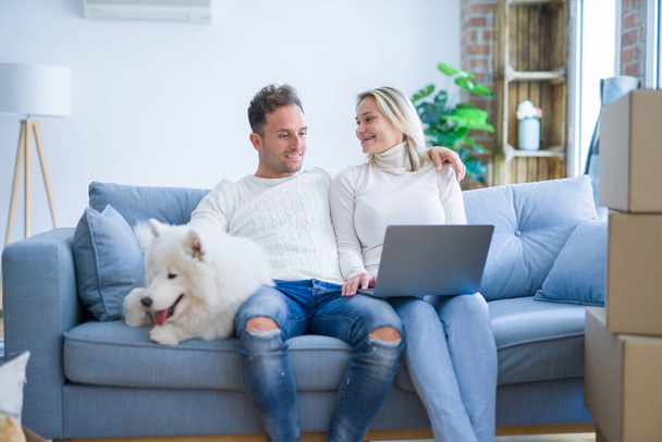 Joven hermosa pareja con perro sentado en el sofá utilizando el ordenador portátil en el nuevo hogar alrededor de cajas de cartón
 - Foto, imagen