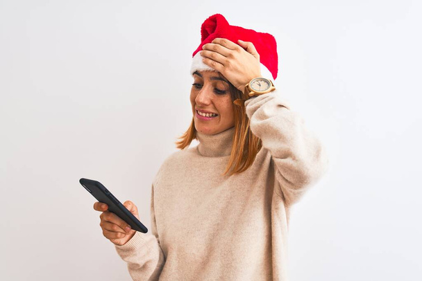 クリスマスの帽子をかぶった美しい赤毛の女性と頭の上に手で強調スマートフォンを使用して、恥と驚きの顔でショックを受け、怒りと不満。恐怖と過ちへの動揺. - 写真・画像