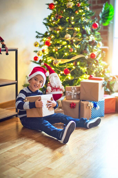 entzückendes Kleinkind, das glücklich und zuversichtlich lächelt. auf dem Boden sitzend mit Weihnachtsmann-Mütze mit Lächeln im Gesicht, Geschenk um den Weihnachtsbaum zu Hause haltend - Foto, Bild