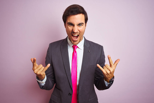 Young handsome businessman wearing suit and tie standing over isolated pink background shouting with crazy expression doing rock symbol with hands up. Music star. Heavy concept. - Foto, immagini