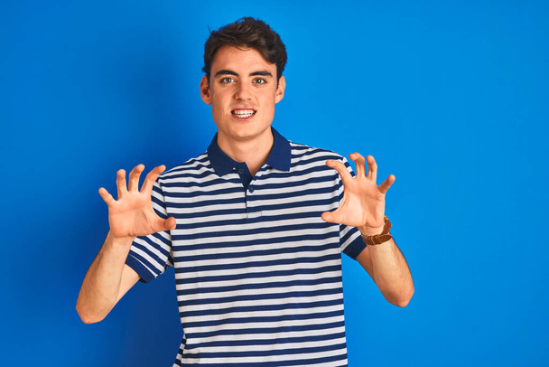 Teenager boy wearing casual t-shirt standing over blue isolated background smiling funny doing claw gesture as cat, aggressive and sexy expression - Φωτογραφία, εικόνα