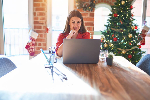 Beautiful woman sitting at the table working with laptop at home around christmas tree looking confident at the camera smiling with crossed arms and hand raised on chin. Thinking positive. - 写真・画像