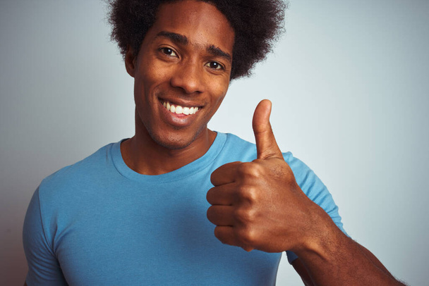 Hombre afroamericano con pelo afro vistiendo camiseta azul de pie sobre fondo blanco aislado feliz con una gran sonrisa haciendo signo ok, pulgar hacia arriba con los dedos, señal excelente
 - Foto, imagen