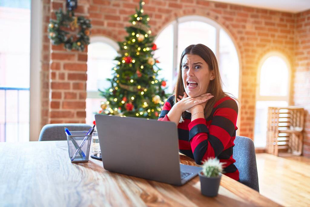Hermosa mujer sentada en la mesa trabajando con el ordenador portátil en casa alrededor de árbol de Navidad gritando y sofocar porque estrangulamiento doloroso. Problema de salud. Concepto de asfixia y suicidio
. - Foto, imagen