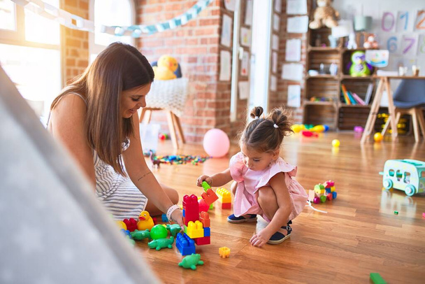 Joven hermosa maestra y niño pequeño sentado en el suelo jugando con bloques de construcción de juguete en el jardín de infantes
 - Foto, Imagen