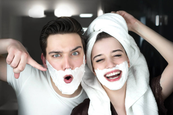young funny couple handsome man and beautiful woman in turban having fun both with shaving foam on their faces in bathroom grimacing playful mood - Fotografie, Obrázek