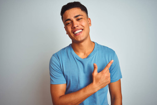 Mladý brazilský muž v modrém tričku stojí nad izolovaným bílým pozadím veselý s úsměvem tváře ukazující rukou a prstem na stranu se šťastným a přirozeným výrazem na tváři - Fotografie, Obrázek