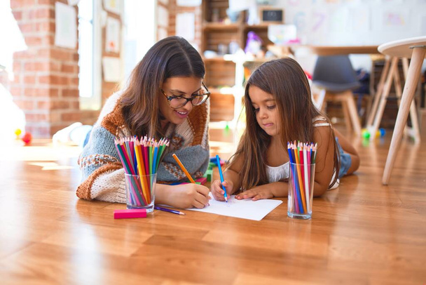 Piękny nauczyciel i maluch rysunek przy użyciu ołówków i papieru wokół wielu zabawek w przedszkolu - Zdjęcie, obraz