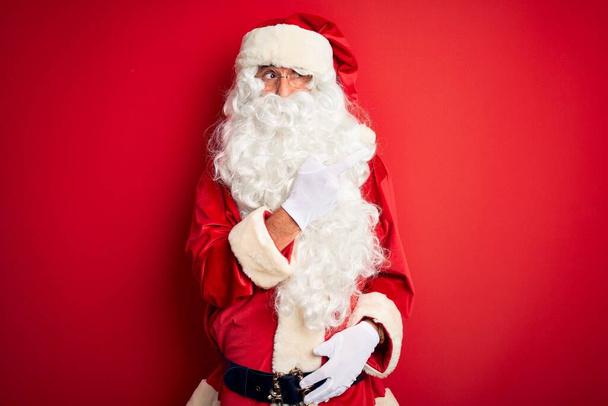 Middle age handsome man wearing Santa costume standing over isolated red background Pointing aside worried and nervous with forefinger, concerned and surprised expression - Photo, Image
