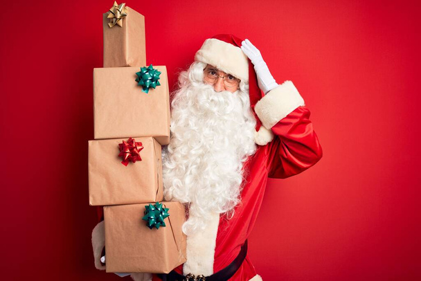 Hombre mayor con traje de Papá Noel sosteniendo torre de regalos sobre fondo rojo aislado estresado con la mano en la cabeza, sorprendido con la vergüenza y la cara sorpresa, enojado y frustrado. Miedo y malestar por error
. - Foto, imagen
