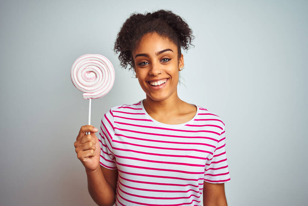Afričanky americká teenager žena jíst barevné cukroví přes izolované bílé pozadí s šťastnou tváří stojící a usmívající se s sebevědomým úsměvem ukazující zuby - Fotografie, Obrázek