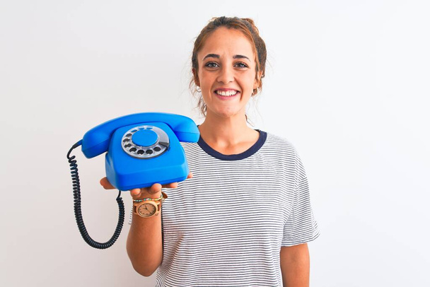Jeune femme rousse tenant un téléphone rétro classique sur fond isolé avec un visage heureux debout et souriant avec un sourire confiant montrant les dents
 - Photo, image