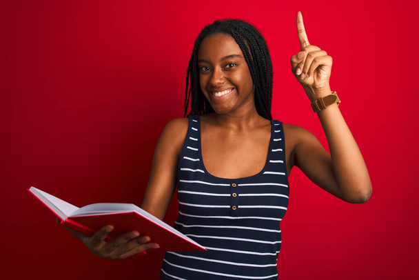 junge afrikanisch-amerikanische Studentin liest Buch steht über isolierten roten Hintergrund überrascht mit einer Idee oder Frage Zeigefinger mit glücklichem Gesicht, Nummer eins - Foto, Bild