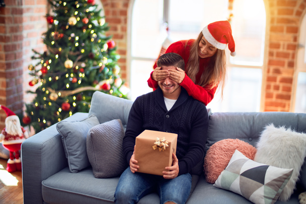 Mutlu ve kendinden emin gülümseyen genç bir çift. Erkeğin gözlerini kapayan bir kadın Noel ağacının etrafında hediyeyle dolaşır. - Fotoğraf, Görsel