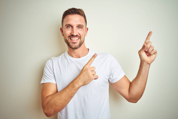 Mladý pohledný muž nosí ležérní bílé tričko izolované pozadí s úsměvem a při pohledu na fotoaparát s oběma rukama a prsty přejdete na stranu. - Fotografie, Obrázek