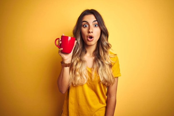 Young beautiful woman holding  red cup of coffee over yellow isolated background scared in shock with a surprise face, afraid and excited with fear expression - Foto, Imagem